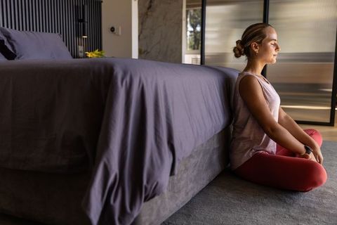 Woman meditating beside bed in minimalist bedroom wearing smartwatch and yoga leggings