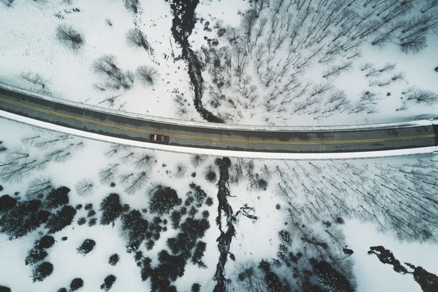 A winter landscape showing a bridge over a creek surrounded by snow-covered trees. The view is from above, captured by a drone. Ideal for use in travel blogs, winter landscape photography, outdoor adventure promotions, nature conservation projects, or seasonal travel guides.