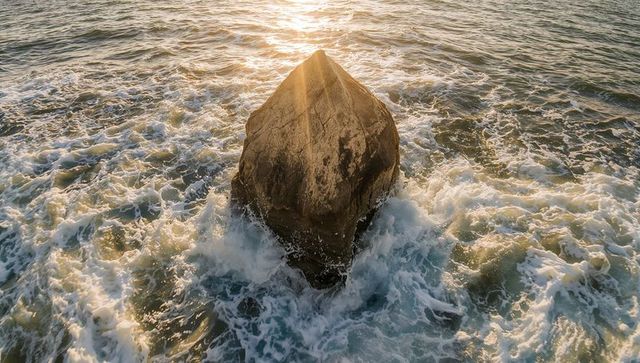 Backlit triangular sea boulder glowing in golden hour waves and foamy surf
