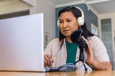 Mature woman recording podcast at home with white headphones and desktop microphone