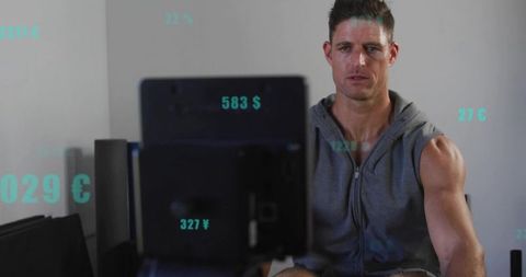 Muscular man in sleeveless hoodie viewing monitor with teal data overlays in modern gym