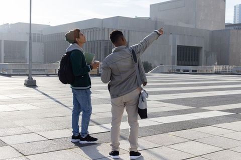 Two diverse young men standing and pointing on urban plaza with backpacks and coffee