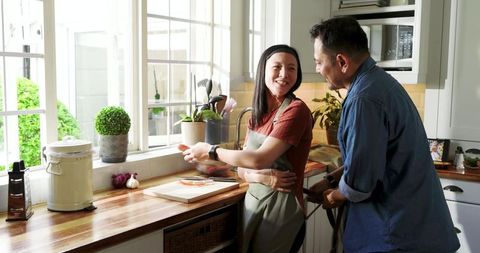 Sunlit asian couple cooking together in cozy kitchen, laughing while chopping vegetables