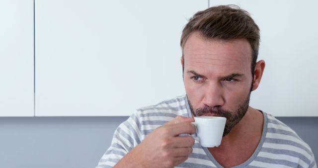 Man drinking espresso, dressed casually in striped shirt. Ideal for use in content focused on morning routines, relaxed home environment, casual lifestyle, and coffee enjoyment.