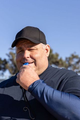 Veteran coach squinting and blowing whistle while signaling team on sunny sports field