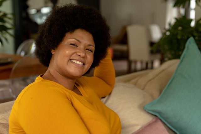 This image depicts a smiling African American mid adult woman sitting comfortably on a sofa at home. Ideal for use in lifestyle blogs, articles on home comfort, mental well-being, and advertisements for home decor or furniture.