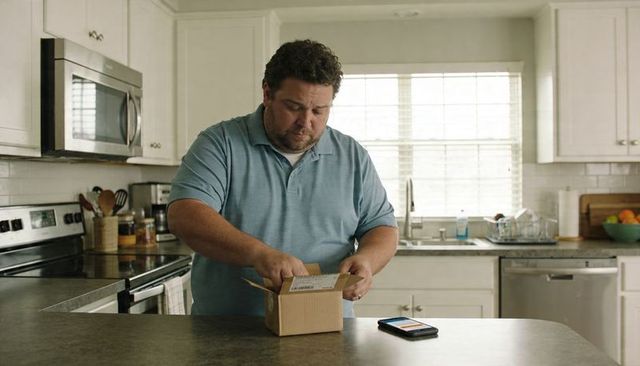 Man Opening Small Cardboard Package on Kitchen Island, Smartphone Displaying Delivery App