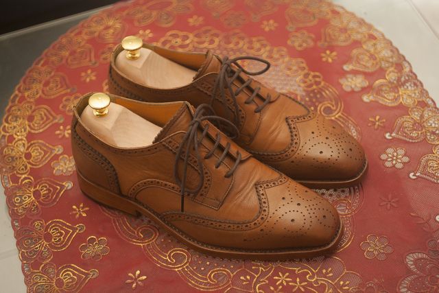 Pair of elegant brown leather dress shoes displayed on intricate red embroidered tablecloth. Ideal for fashion stores, formal wear promotions, men's accessories advertising, vintage fashion collections, or luxury footwear features.