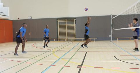 Diverse male volleyball team jumping and passing ball over net on indoor gym court