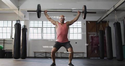 Male Athlete Lifting Olympic Barbell Overhead in Gym with Hanging Punching Bags