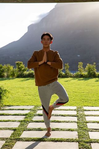 Asian man practicing tree pose on stepping stones balancing barefoot in garden at sunrise