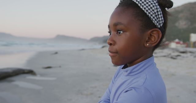 An African American girl stands at the beach, gazing at the horizon with a serene expression. She wears a checkered headband and a light purple top. This image can be used for topics on childhood, contemplation, nature's beauty, and peaceful moments. Ideal for use in blogs, educational content, and promotional material promoting relaxation and enjoying time outdoors.