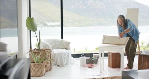 Mature artist arranging stretched canvas in bright coastal studio with ocean view