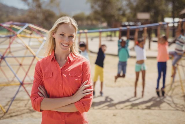Female teacher standing with arms crossed, smiling confidently at school playground. Children are playing in the background, enjoying outdoor activities. Ideal for use in educational materials, school brochures, teacher recruitment ads, and articles about education and child development.