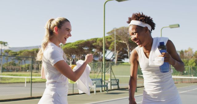 Two female friends happily engaging after a tennis match on an outdoor court. One is holding a towel and the other a water bottle, both dressed in tennis attire, representing fitness, exercise, and an active lifestyle. Perfect for promotions, fitness campaigns, team-building activities, sports equipment, and outdoor recreational topics.