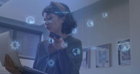 Asian woman holding laptop and speaking into phone with holographic AR icons, home office