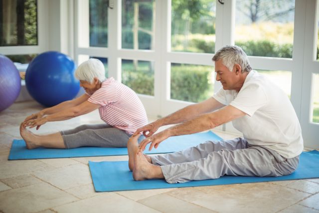 Senior couple engaging in stretching exercises on blue exercise mats in a bright, spacious room with large windows. Ideal for use in articles or advertisements promoting senior fitness, healthy lifestyles for elderly, home workout routines, and wellness programs for mature adults.