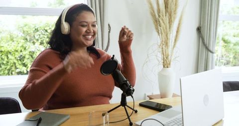 Smiling woman recording podcast at home workspace with headset, microphone, laptop, smartphone