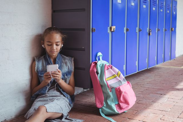 Elementary student listening music through headphones while using mobile phone by lockers at school