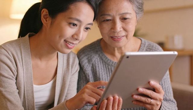 Younger woman teaching senior tablet skills at home, multigenerational family learning together