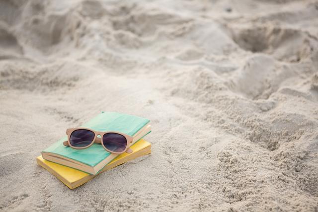Sunglasses resting on two books on sandy beach, evoking feelings of summer relaxation and leisure. Ideal for use in travel blogs, vacation advertisements, lifestyle articles, and summer-themed promotions.