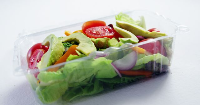 A fresh vegetable salad in a clear plastic container sits on a white surface, with copy space. The salad includes a variety of colorful ingredients like lettuce, tomatoes, and avocado, emphasizing a healthy eating choice.