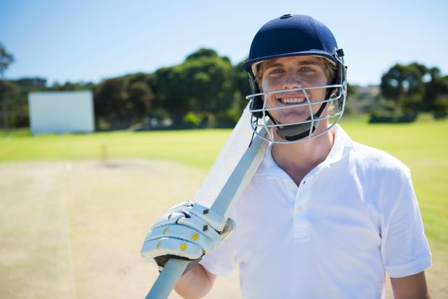 Portrait of smiling cricket player holding bat while wearing helmet at field on sunny day