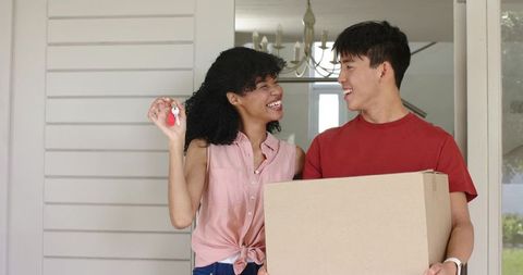 Smiling diverse couple moving into new home holding keys and carrying moving box