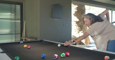 Leaning woman aiming cue on black-felt pool table in modern home gameroom, senior leisure