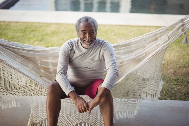 Senior man sitting on hammock and smiling. Ideal for illustrating themes of relaxation, outdoor leisure, healthy aging, retirement lifestyle, and happiness. Suitable for lifestyle blogs, retirement planning materials, and health and wellness content.