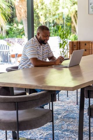 African man working from home at dining table with laptop and garden view, natural light