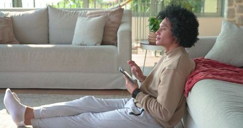 Mid-adult african american man sitting on rug by sofa scrolling tablet and relaxing at home
