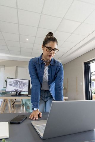 Female architect leaning over laptop reviewing 3d floorplan on monitor in modern studio
