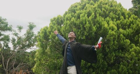 Male graduate celebrating outdoors holding diploma with red ribbon near evergreen trees
