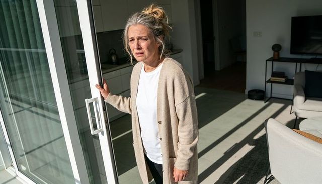 Worried senior woman holding sliding glass door handle in sunlit modern apartment balcony