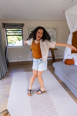 Joyful african american woman dancing in bright bedroom wearing rust tank top and denim shorts
