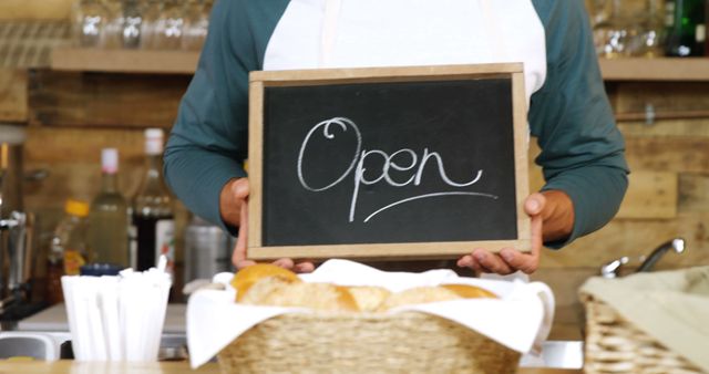 Barista holding chalkboard with the word 'Open' in cozy coffee shop setting. Baskets with bread blurred in foreground. Could be used for promoting small businesses, bakeries, coffee shops, opening announcements.