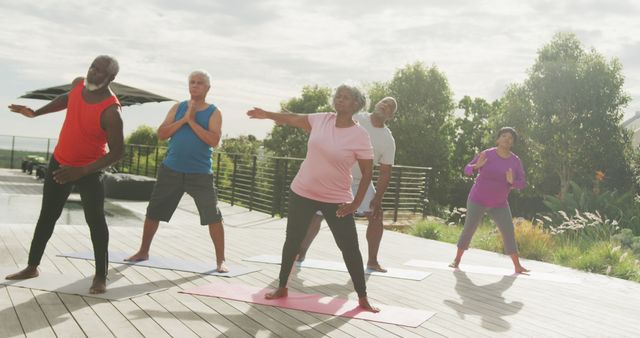 Group of senior individuals practicing yoga outdoors on a wooden deck surrounded by lush greenery. They are engaged in various yoga poses, showcasing their dedication to maintaining a healthy lifestyle. Useful for themes related to senior fitness, outdoor activities, wellness programs, and healthy aging. Ideal for use in health blogs, fitness programs aimed at older adults, and advertisements promoting an active lifestyle for seniors.