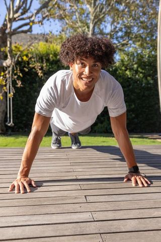 Smiling young man performing outdoor push-ups on wooden deck during sunny backyard workout
