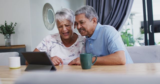 Senior couple sitting at table, using tablet for video calling. Ideal for topics on senior technology adoption, family connections, and senior lifestyle.