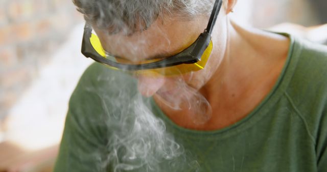 Person wearing yellow-tinted safety glasses appearing focused while working with smoke rising. Useful for illustrating workplace safety, protective gear usage, hazardous environments, and industrial work conditions.
