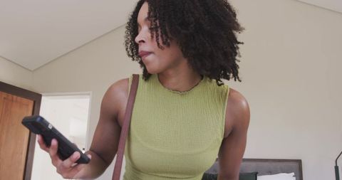 Adult african woman holding smartphone, looking left in modern bedroom with green top