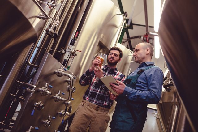 Worker and owner in a brewery discussing beer quality while examining a sample. Ideal for use in articles about the brewing process, quality control in beverage production, teamwork in industrial settings, and the craft beer industry.