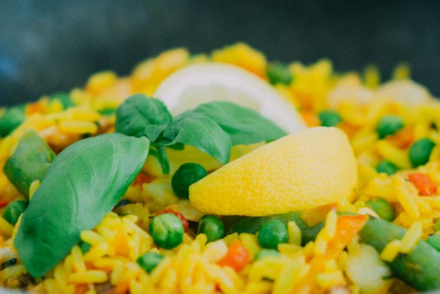 Close-up of colorful vegetarian paella featuring fresh basil leaves and a slice of lemon. The vibrant meal includes green peas, red bell peppers, and yellow rice, creating a visually appealing dish. Ideal for use in food-related articles, restaurant menus, culinary blogs, healthy eating promotions, and Mediterranean cuisine features.