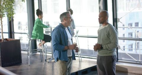Multiracial coworkers chatting over coffee in modern glass office lounge with city view