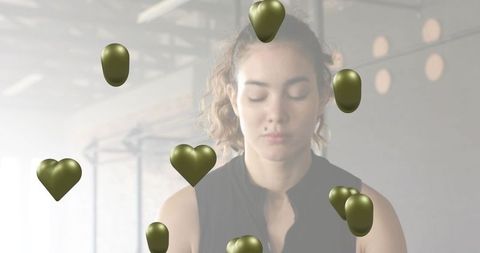 Young woman meditating with floating olive-gold heart shapes in industrial studio for mindfulness