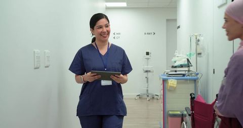 Nurse holding tablet speaking to woman in wheelchair in hospital corridor compassionate care