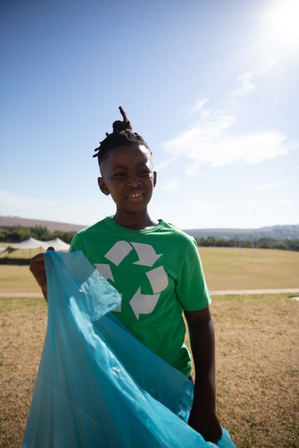 Young boy wearing a green recycling shirt holding a blue trash bag in a park on a sunny day. Ideal for promoting environmental awareness, sustainability education, community clean-up events, and eco-friendly initiatives. Perfect for use in educational materials, environmental campaigns, and social responsibility projects.
