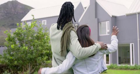 Diverse couple embracing on lawn in front of modern gray suburban house