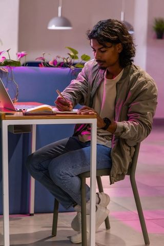 Young indian man writing in notebook at cozy coworking lounge with laptop and plants